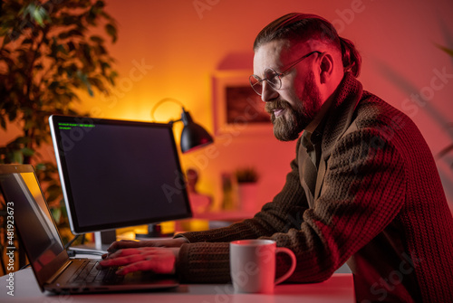 Positive bearded man in eyeglasses using modern computers for writing data code during night at home. IT specialist working late at cozy home.