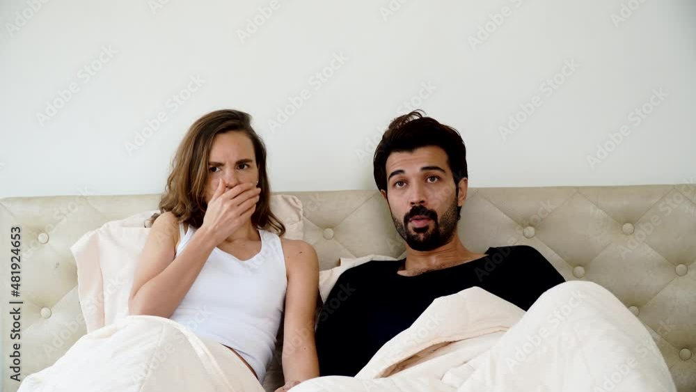 Scared young couple sitting under blanket on the bed with laptop while watching horror movie at tv together in the bedroom. Husband embarrassed his wife while looking scary shocked at home. Scared young couple sitting under blanket on the bed with laptop while watching horror movie at tv together in the bedroom. Husband embarrassed his wife while looking scary shocked at home.