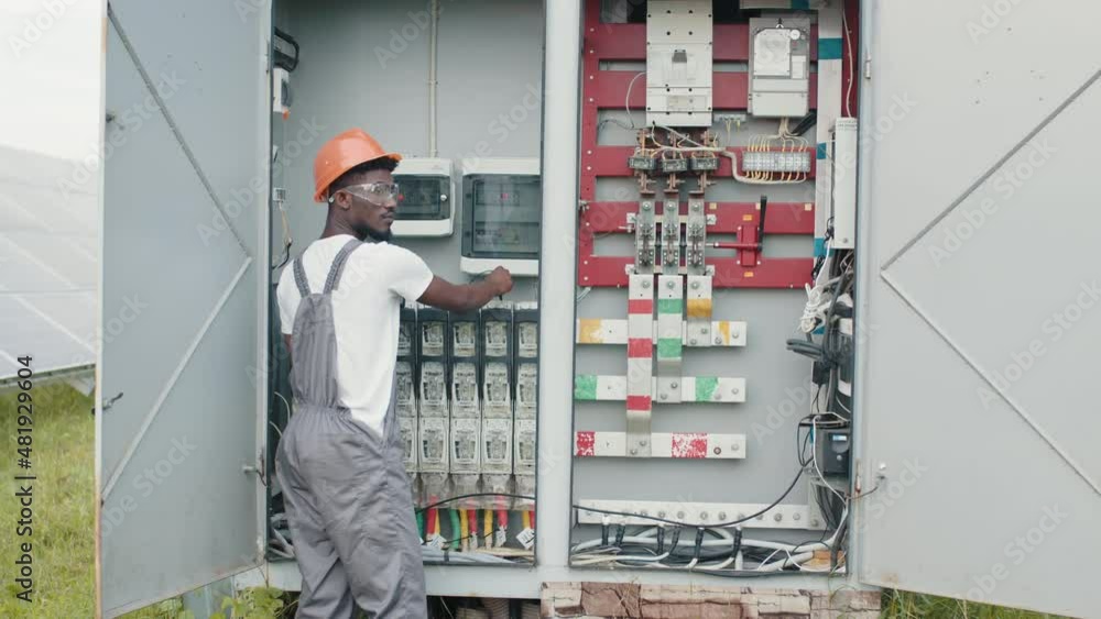 African american man in grey overalls and orange helmet repairing cables in switchgear at solar station. Concept of people, maintenance and alternative energy African american man in grey overalls and orange helmet repairing cables in switchgear at solar station. Concept of people, maintenance and alternative energy