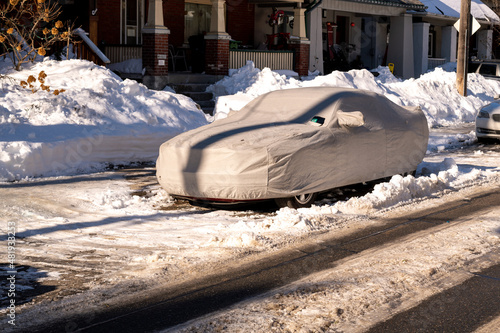 A car sits parked on a snow filled residential street protected by a fabric car cover.