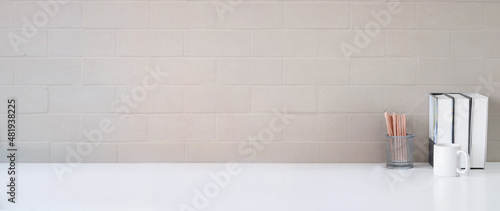 Cup of coffee, books and pencil holder on white table with brick wall. Copy space for your advertising text.