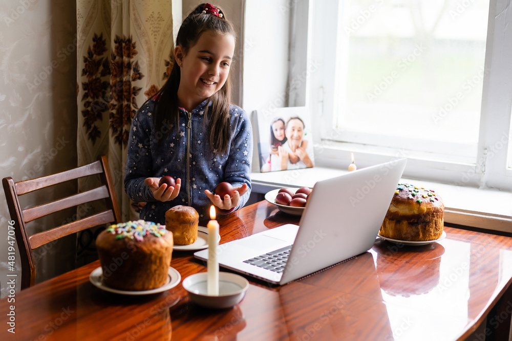 Pretty little girl painting easter eggs at home during coronavirus covid-19 outbreak. Kid using laptop on kitchen, online, video call to friends