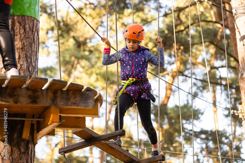 Little girl is standing on a rope, holding a rope with his hands. A child in a rope park passes obstacles