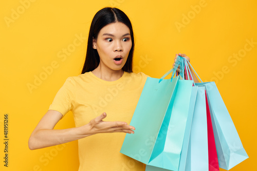 Charming young Asian woman in a yellow T-shirt with multicolored shopping bags yellow background unaltered