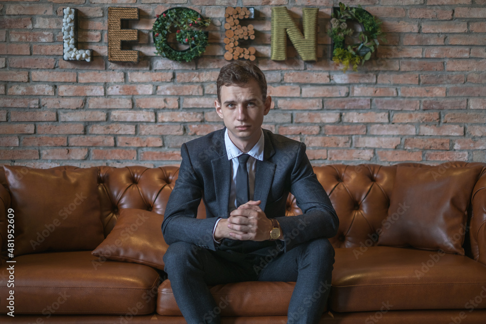 Portrait of young caucasian business man in formal suit sitting on brown leather sofa. Handsome manager sitting on a couch in an open space in office Portrait of young caucasian business man in formal suit sitting on brown leather sofa. Handsome manager sitting on a couch in an open space in office