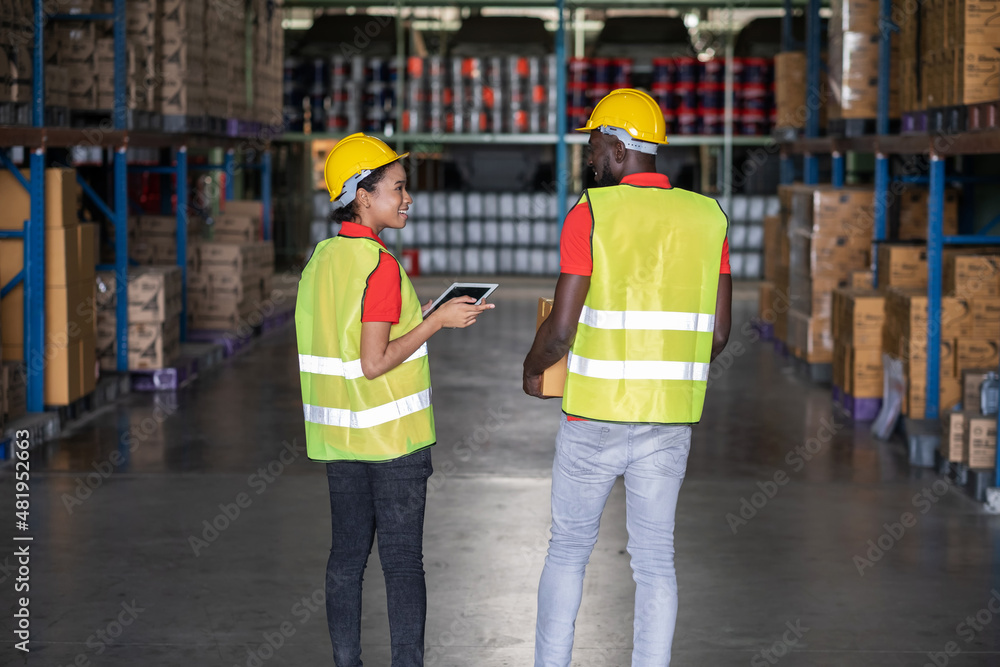 Back view of Africa American engineer woman holding tablet wearing safety helmet and vest standing in the automotive part warehouse. Looking at friend. Portrait of worker. Logistic and business. Back view of Africa American engineer woman holding tablet wearing safety helmet and vest standing in the automotive part warehouse. Looking at friend. Portrait of worker. Logistic and business.