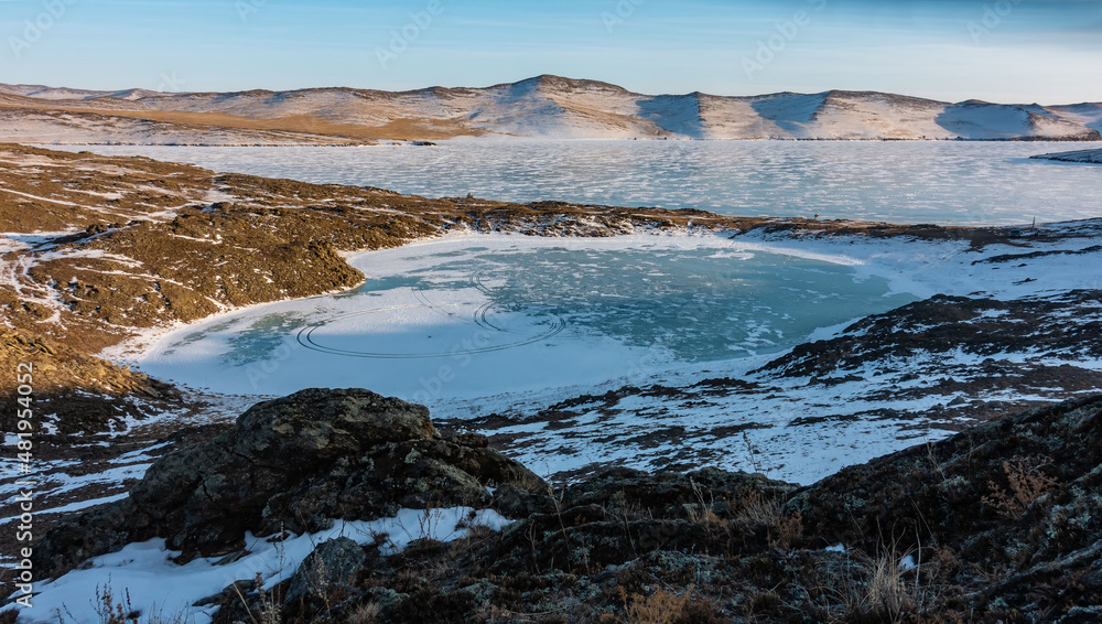 An amazing little frozen lake in the shape of a heart. Tire tracks are visible on the turquoise ice. Snow on the ground. A mountain range against a blue sky. Big Lake Baikal is nearby. An amazing little frozen lake in the shape of a heart. Tire tracks are visible on the turquoise ice. Snow on the ground. A mountain range against a blue sky. Big Lake Baikal is nearby.