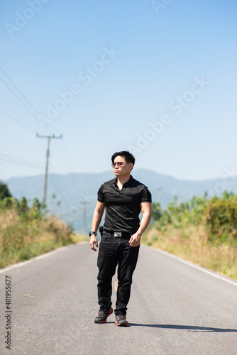 thai asian man short hair serious face wear black shirt plat wrist watch and shade walking with confident bollywood style on middle of asphalt street and mountain in background