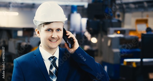 Portrait of a professional heavy industry engineer in protective uniform and hard hat, smiling charmingly. Unfocused large industrial plant in the background