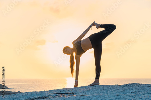 Young woman with long hair in a  black sportswear doing yoga exercises on the rocks by the sea. Sunrise sky on the background.
