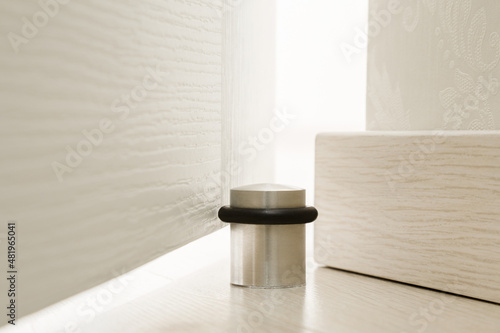 Stainless door stopper with black rubber behind white door on light wooden floor. Protection for wall, wallpaper or baseboard against damages of door bump. Front view. Closeup. photo