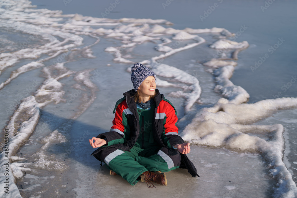 A woman in a colorful thermal costume enjoying winter while sitting in a meditation pose on a frozen lake with blue sky in the background. Freedom, peace, quiet, serenity A woman in a colorful thermal costume enjoying winter while sitting in a meditation pose on a frozen lake with blue sky in the background. Freedom, peace, quiet, serenity