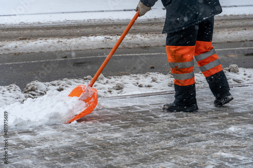 Removing snow from the sidewalk after snowstorm. A road worker with a shovel in his hands and in special clothes cleans the sidewalk and the road from snow. Snowstorm and hurricane in the city. © Pokoman