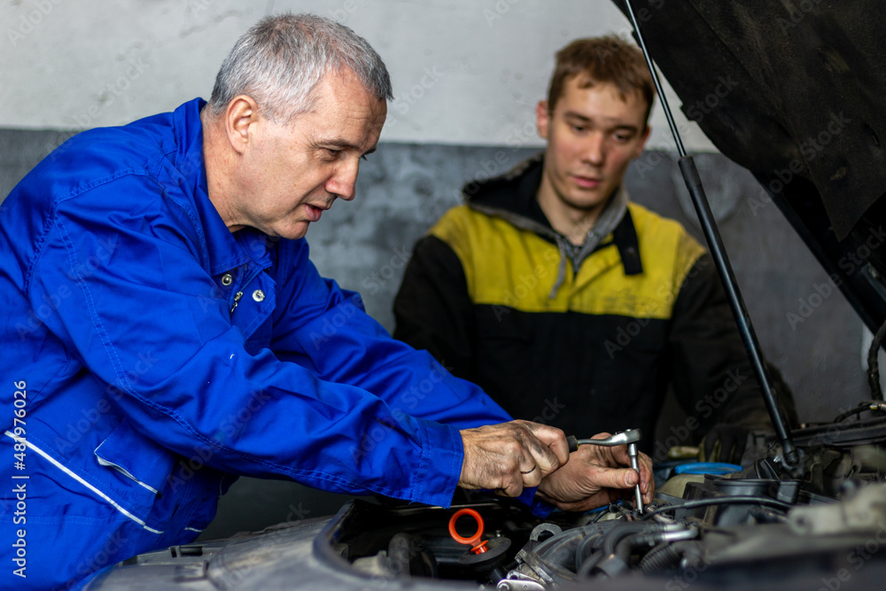 Senior experienced mechanic teaches his colleague how to repair a car