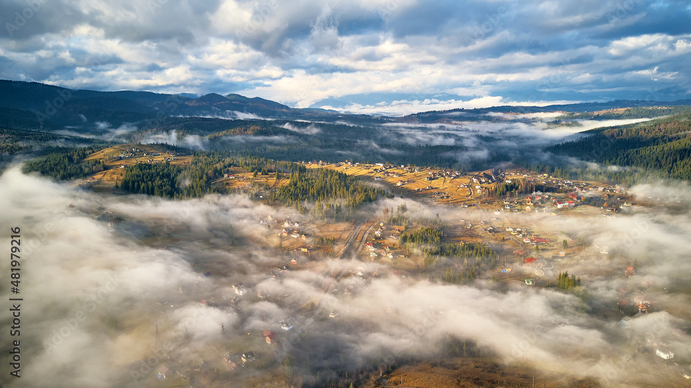 Colorful autumn morning over foggy mountain valley. Village covered by low clouds. Fall rural landscape. Misty cold November morning.