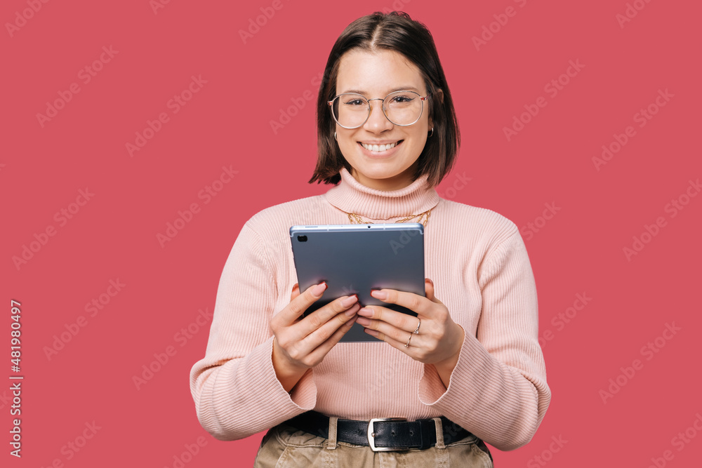 Young joyful woman using tablet over pink background and looking confident at the camera. Young joyful woman using tablet over pink background and looking confident at the camera.