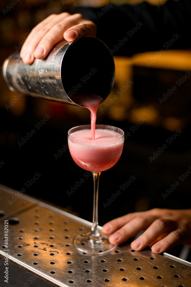 hand of the bartender holds shaker and pours pink cocktail into glass