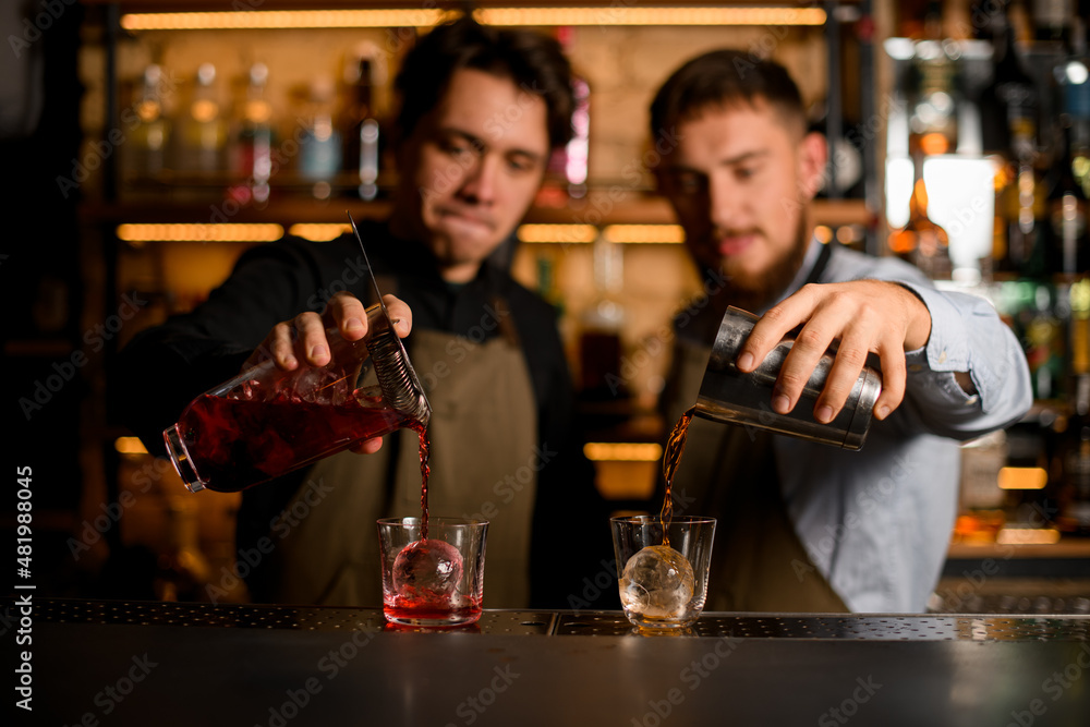 two glasses with ice in which bartenders carefully pour different cocktails