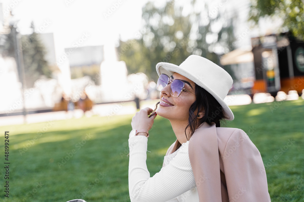 Lifestyle portrait of fashion girl weared white hat, dress, jacket and sunglasses . Woman is sitting on the grass.
