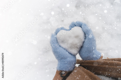 Cozy female hands in blue fluffy mitten with heart of snow in winter day. Love concept. Snow falls. Valentine day background. Copy space for design.