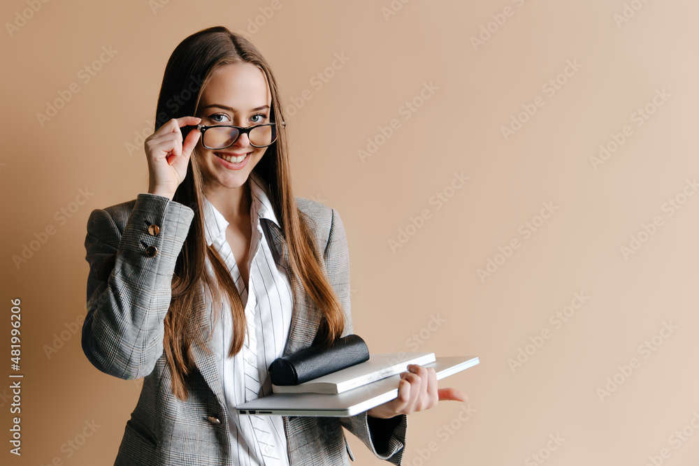 Young woman is touching her glasses, holding notebook and laptop, looking at the camera isolated on beige background. Businesswoman talking to her colleague Young woman is touching her glasses, holding notebook and laptop, looking at the camera isolated on beige background. Businesswoman talking to her colleague