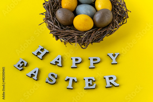 Top view of multicolored Easter eggs in birds nest on yellow background. Happy easter lettering.