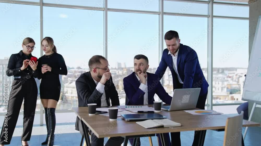 Men in office talking about work sitting at the desk with papers and laptop. Business ladies standing at the backdrop. Panoramic windows with city view at the backdrop. Men in office talking about work sitting at the desk with papers and laptop. Business ladies standing at the backdrop. Panoramic windows with city view at the backdrop.