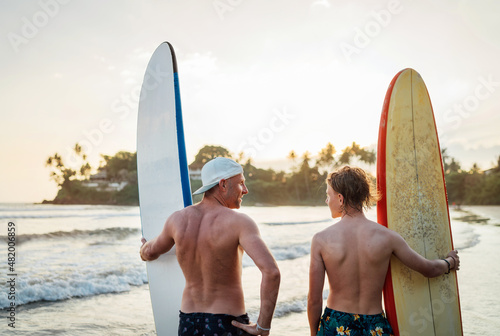Father with teenager son standing with surfboards on the sandy ocean beach with palm trees on background lightened with sunset sun. They smiling and have a conversation. Family active vacation concept © Soloviova Liudmyla