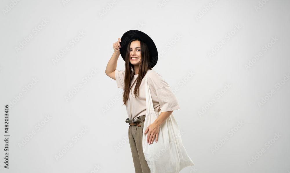 Brunette woman in a beige t-shirt and black hat with a cotton shopper bag with groceries looking in a camera. Reusable eco bag for shopping. Zero waste concept.