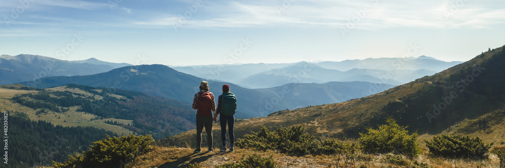 Couple of hikers with backpacks enjoying valley landscape view from top of a mountain. Young adult tourists, man and woman standing on the pass and holding by hands. Panoramic view of mountain hills