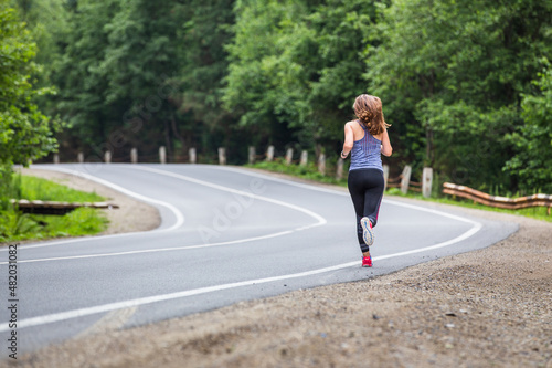 Young fitness woman running on the road through the forest. Slim girl jogging in the morning along the road.
