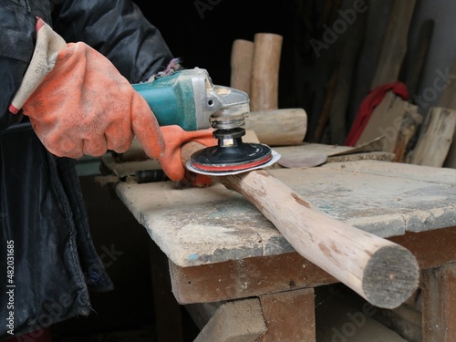 smoothing a thin log with a grinder with a sandpaper nozzle, carpenter's hands in gloves with an angle saw for processing wooden parts on a carpentry work surface, a grinder with an emery nozzle