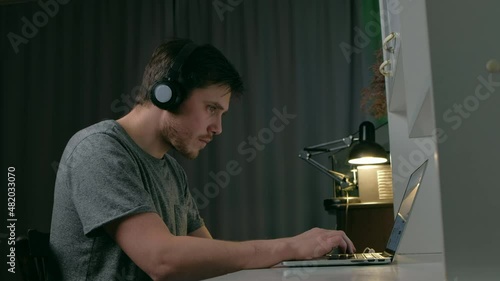 Male on headphones in slouching position sitting listening music on laptop sits at table at night in home or office. Working late use computer. Concept of distance work during the coronavirus pandemic photo