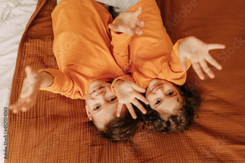 Smiling cheerful children a little girl and a boy in bright orange pajamas hug and lie resting on the bed in a cozy bedroom at home on a weekend indoor. Top view. Selective focus
