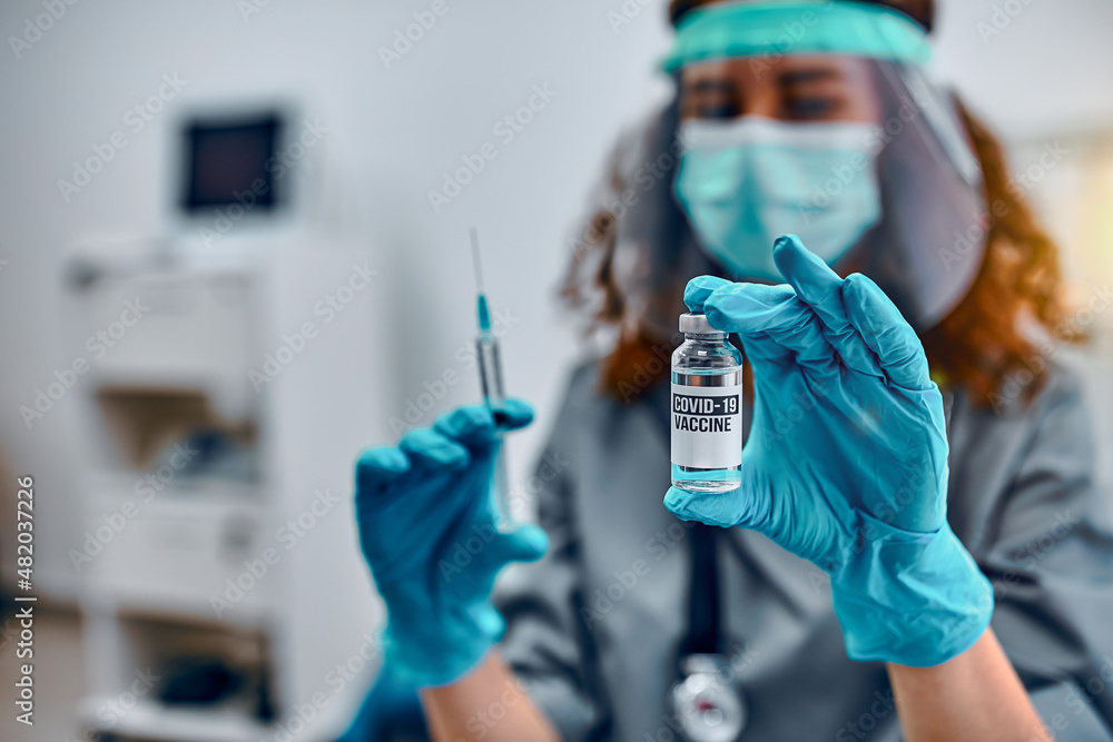 Vaccination and preservation and protection of health. A female doctor in a protective mask and protective medical gloves holds a covid-19 vaccine and a syringe. Vaccination and preservation and protection of health. A female doctor in a protective mask and protective medical gloves holds a covid-19 vaccine and a syringe.
