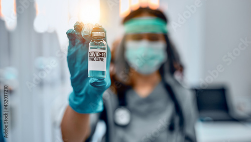 A female doctor in a protective mask and protective medical gloves holds a bottle of Vaccine-19 vaccine, a close-up image and inscription. Vaccination and preservation and protection of health.