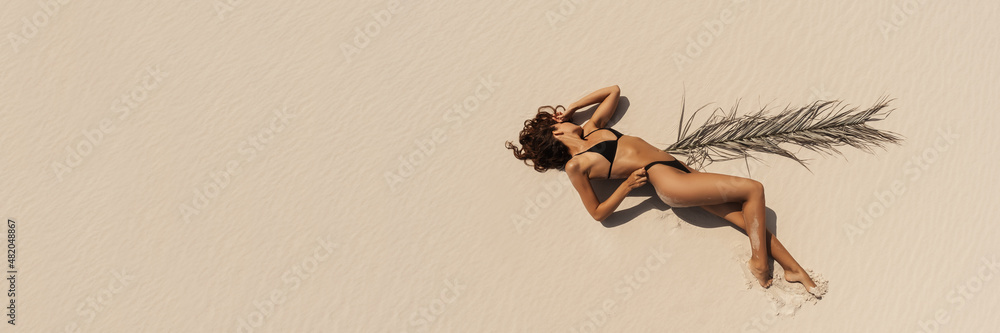 Top aerial drone view of woman in swimsuit relaxing and sunbathing on beach Near The Ocean. Attractive brunette girl in black bikini laying on a sandy coast with palm leaf. Gorgeous tanned mixed race - obrazy, fototapety, plakaty 