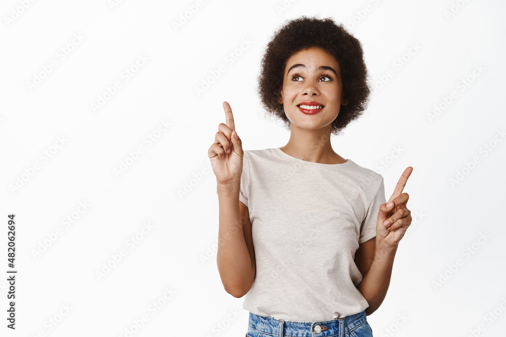 Portrait of smiling black woman pointing fingers up and looking at top, advertisement, reading promo text copyspace, standing over white background Portrait of smiling black woman pointing fingers up and looking at top, advertisement, reading promo text copyspace, standing over white background