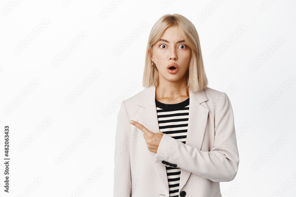 Image of businesswoman with surprised face, pointing finger left at empty copy space, looking shocked, say wow, staring at camera impressed, white background Image of businesswoman with surprised face, pointing finger left at empty copy space, looking shocked, say wow, staring at camera impressed, white background