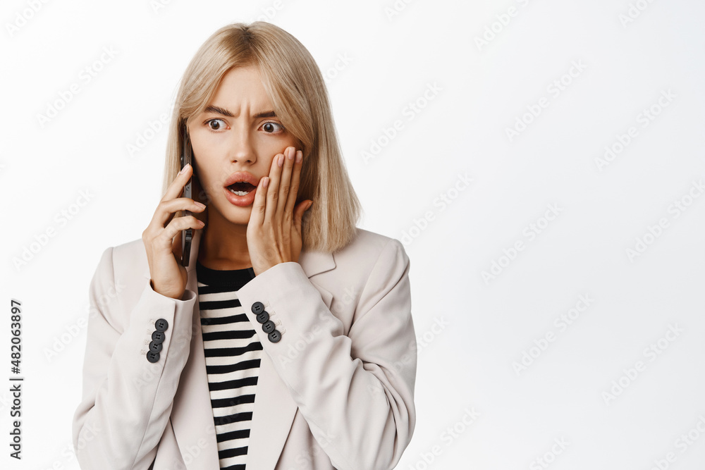 Image of young woman reacting shocked to phone call, standing concerned against white studio background Image of young woman reacting shocked to phone call, standing concerned against white studio background