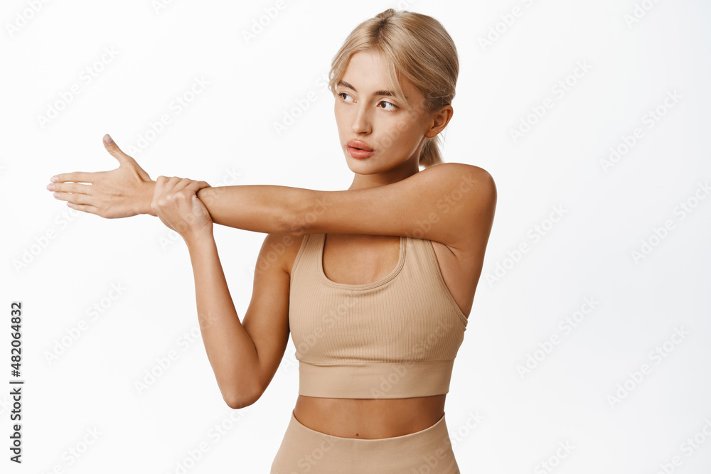Portrait of female athlete, sportswoman in sport bra and leggings, warm up for workout, fitness or yoga, stretching arms, doing hand exercises, white background Portrait of female athlete, sportswoman in sport bra and leggings, warm up for workout, fitness or yoga, stretching arms, doing hand exercises, white background