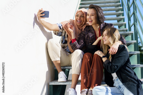 Happy multiethnic female best friends taking selfie sitting on stairs in