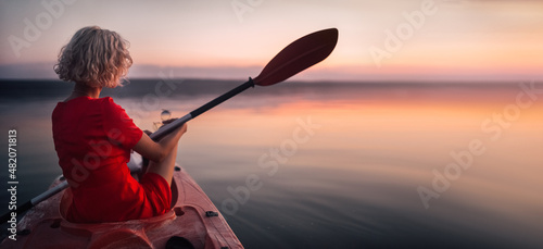 Blonde woman in a red dress sits in a boat with a paddle at sunset.