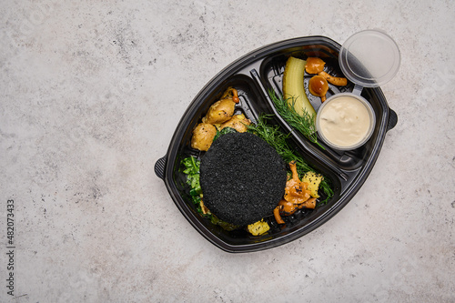 Rabbit cutlet in black breading in baby potatoes, spinach, sauce, and mushrooms in black plastic container on light background flat lay. Designer food concept for take away or delivery