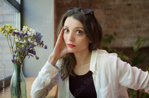 Indoor female portrait of a thoughtful girl in black top and white shirt sitting near the window in loft style cafe