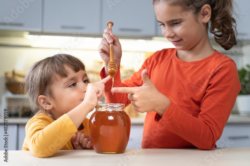 Happy children eating honey from a jar at home in the kitchen. funny girls lick honey from a spoon. Healthy organic food sweets for kids