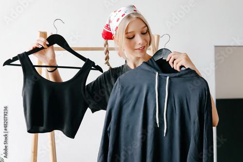 Young lady chossing what to buy, holding hangers with top and hoodie in designer store photo