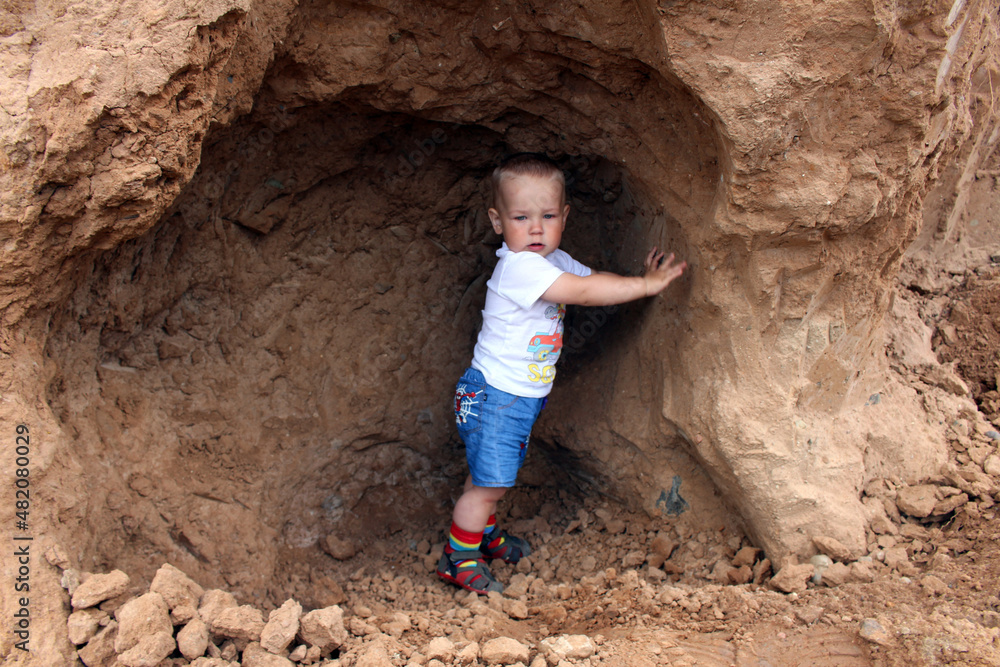 A little boy with blond hair on a walk outside on a summer day. The boy is sitting in a clay cave.