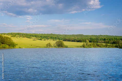 Lake, pond, on the other side of the field and green forest. Charming nature. Landscape with beautiful sky and river.