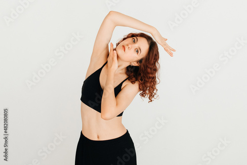 Cheerful woman with curly red hair smiling  posing with her hand near her face in black sport top  standing at white background